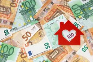 Red felt small house on right side of 50 and 100 euro banknotes background. Heart-shaped hole is cut instead of a window. Real estate loan concept. Money for a dream house. Top view. Place for text. photo