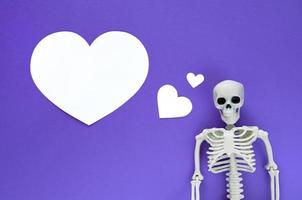 Skeleton on violet background with white blank paper cut hearts, one big heart with copy space. Anatomical plastic model human skeleton with dreaming text balloon. Valentine's day love concept.