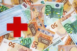 KRAKOW, POLAND, MARCH 10, 2021 - Medical cross symbol cut out of red felt with part of blue medical mask on background with 50 and 100 euro banknotes. Healthcare system in Europe. Paid medicine. photo