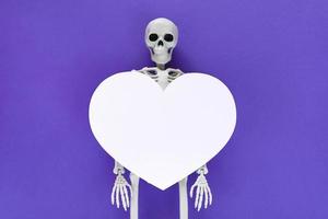 Skeleton with big white blank cut out heart on it, place for text in paper heart frame. Anatomical plastic model human skeleton centered on violet background. Valentine's day love concept.