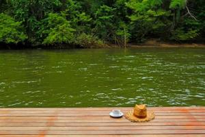 Take a relax time with cup of coffee and straw hat on brown wooden floor with blurred river and green forest background on vacation day at Kanchanaburi, Thailand- Beauty of Nature and Landmark concept photo