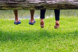 cerca arriba piernas de Pareja en amor sentado juntos en madera o madera banco y césped piso con lago ver antecedentes. inferior ver cuerpo parte de gente. relajante y amante hora con Copiar espacio. foto