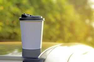 café para llevar en un papel taza en parte superior de el coche techo verde árbol antecedentes a amanecer en el mañana, selectivo enfocar, suave enfocar. foto