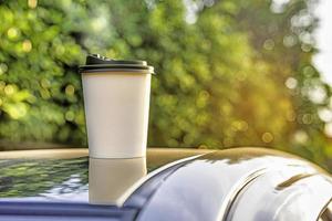 coffee takeaway in a paper cup on top of the car roof green tree background at sunrise in the morning,  selective focus, soft focus. photo