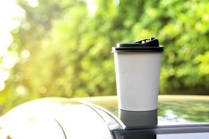 café para llevar en un papel taza en parte superior de el coche techo verde árbol antecedentes a amanecer en el mañana, selectivo enfocar, suave enfocar. foto