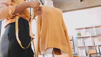 Moda diseñador l joven asiático mujer trabajando utilizando computadora portátil, tableta y sonriente mientras en pie en taller respondiendo en negocio video