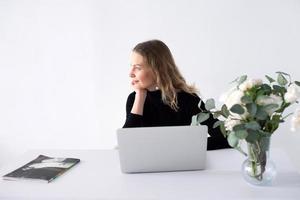 el niña trabajos en un blanco oficina con un ordenador portátil y flores en el mesa foto