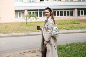 A pretty girl student in a coat with a cup of coffee is walking near the educational institution photo