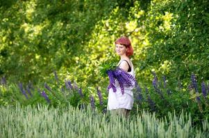 A young girl in a white shirt with pink hair stands in a green field with a bouquet of lupines photo