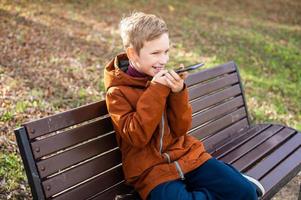 un chico registros un voz mensaje y se ríe mientras sentado en un parque banco en otoño foto