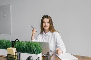 A cute girl is sitting at a desk in the office working with a laptop and notepad and looking at the camera photo