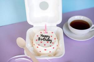 A small bento cake in eco-packaging with a wooden spoon and a candle next to a cup of tea on a purple and blue background photo