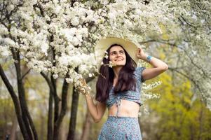 un joven niña en un sombrero y con tulipanes, en pie en un azul vestir siguiente a un floreciente blanco árbol. foto