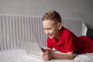 A cute boy in a red T-shirt is lying on the bed with a tablet in his hands photo