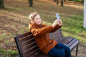 A boy takes a selfie on his phone while sitting on a park bench photo