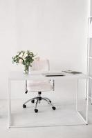 oficina ambiente. un blanco habitación con un escritorio en cuales allí es un florero, un ordenador portátil y revistas foto