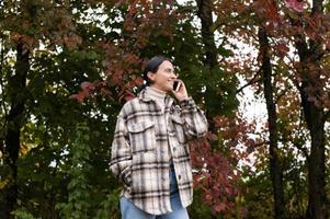 un linda niña tiene un agradable conversacion en el teléfono en contra el antecedentes de un otoño bosque foto