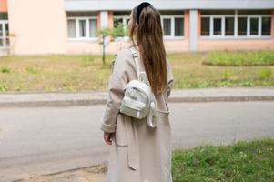 A pretty girl student in a coat and with a white backpack is walking near the educational institution photo