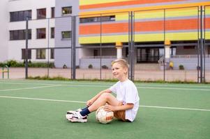 A teenage boy is sitting on a green field in the school yard with a soccer ball photo