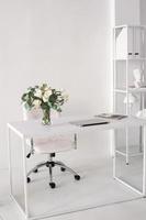 Office environment. A white room with a desk on which there is a vase, a laptop and magazines photo