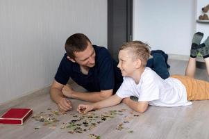 un linda chico con un alegre papá es coleccionar un rompecabezas acostado en el piso en un habitación foto