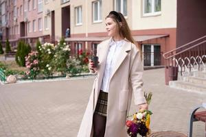 A cute girl in a coat holds a glass of coffee and a bouquet of flowers on the street photo