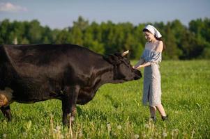 un joven niña en un vaca pasto soportes en un vestir y un pañuelo y golpes un vaca foto