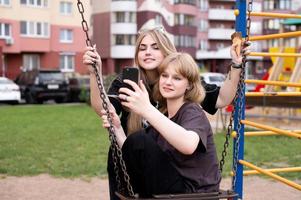 Two funny girls are sitting on a swing in the city and taking selfies on their phone. Teenagers smile photo