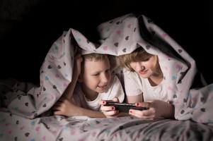 Brother and sister lie under a blanket at night and play on the phone photo