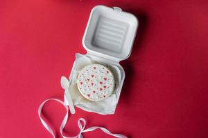 un abierto caja con un San Valentín día bento pastel con corazones en un rojo antecedentes con un blanco cinta foto