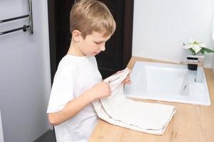 The boy folds a towel and stands in the bathroom near the washbasin photo