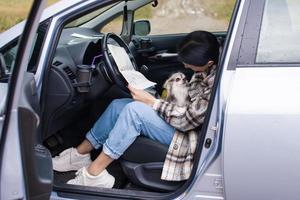 A cute girl is sitting in a car behind the wheel, holding a chihuahua and a road map, smiling photo