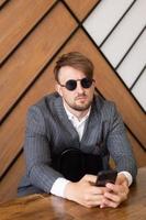 A young man in a suit and sunglasses is sitting at a desk in the office and looking at the phone photo