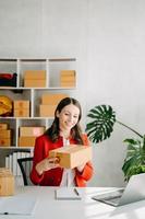 puesta en marcha pequeño negocio pequeño, empresario propietario mujer utilizando teléfono inteligente o tableta tomando recibir y comprobación en línea compra compras orden a preparando paquete producto caja. foto