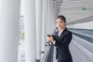 joven asiático mujer de negocios en negro traje utilizando digital tableta y en pie en pasarela estación con Copiar espacio. foto