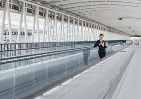 young Asian businesswoman in black suit using phone and holding cup of coffee and standing on elevated walkway station with copy space. photo