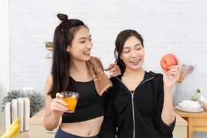 Asian active two women sibling in sportswear enjoy eating fruits healthy foods. Beautiful girl, sister holding a glass of orange juice and apples in the kitchen photo