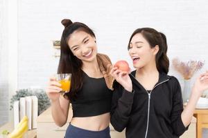 Asian active two women sibling in sportswear enjoy eating fruits healthy foods. Beautiful girl, sister holding a glass of orange juice and apples in the kitchen photo