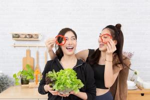 Asian active two women sibling in sportswear enjoy eating fruits healthy foods. Beautiful girl, sister holding Bell Pepper, sliced like glasses. In the kitchen. photo