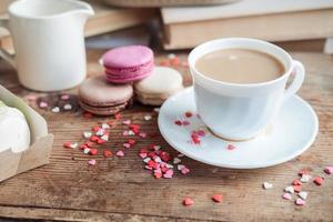 Macaroons and a cup of coffee, a milk jug on a background of small hearts on a wooden background, top view photo
