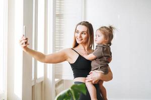Young mother in sportwear with baby girl on hands taking selfie on mobile phone in bright room photo