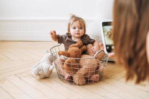 Cute baby girl sitting in basket with soft toys and young mother taking photo on mobile phone at home