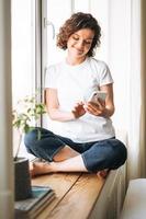 Young brunette woman in casual clothes using mobile phone sitting on window sill at home photo