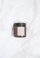 Glass jar with empty label on white sheets, bed linen background, mockup photo