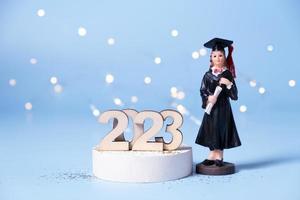 Class of 2023 concept. Wooden number 2023 with graduate statuette on colored background with bokeh photo