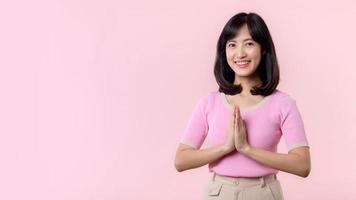 Portrait beautiful young asian woman happy smile with Sawasdee, hello gesture in Thai culture Siam traditional greeting style isolated on pink pastel background. photo