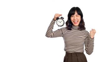 Smiling cheerful attractive young asian woman wearing sweater shirt holding alarm clock showing fist up looking camera isolated on white background, studio portrait photo