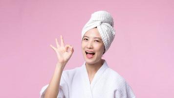 Beautiful young asian woman happy smile face with hand or finger gesture signs in bathrobe and shower towel showing empty space isolated on pink background. photo