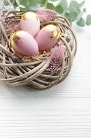 Easter eggs painted pink and gold and  feathers in a nest on a white wooden background.