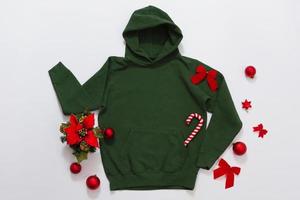 Close up green blank template hoodie copy space. Christmas Holiday concept. Top view mockup hoodie. Red holidays decorations on jumper white background. Happy New Year accessories. Selective focus photo